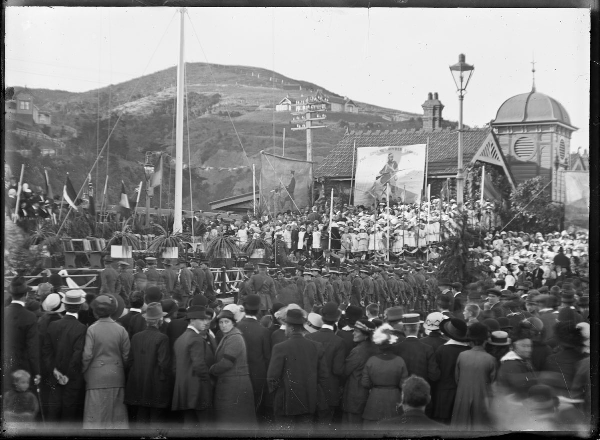 Anzac day commemoration at Petone, on 25 April 1916. Shows a crowd, flagpole and banners next to the Petone Railway Station. 